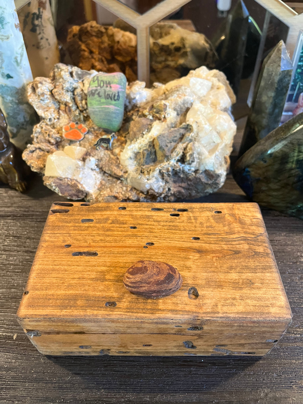 A rustic wooden keepsake box with a lid, featuring a small oval piece of Oregon driftwood on the center and several small cutouts on the lid, displayed on a dark wooden surface with crystals and minerals in the background.