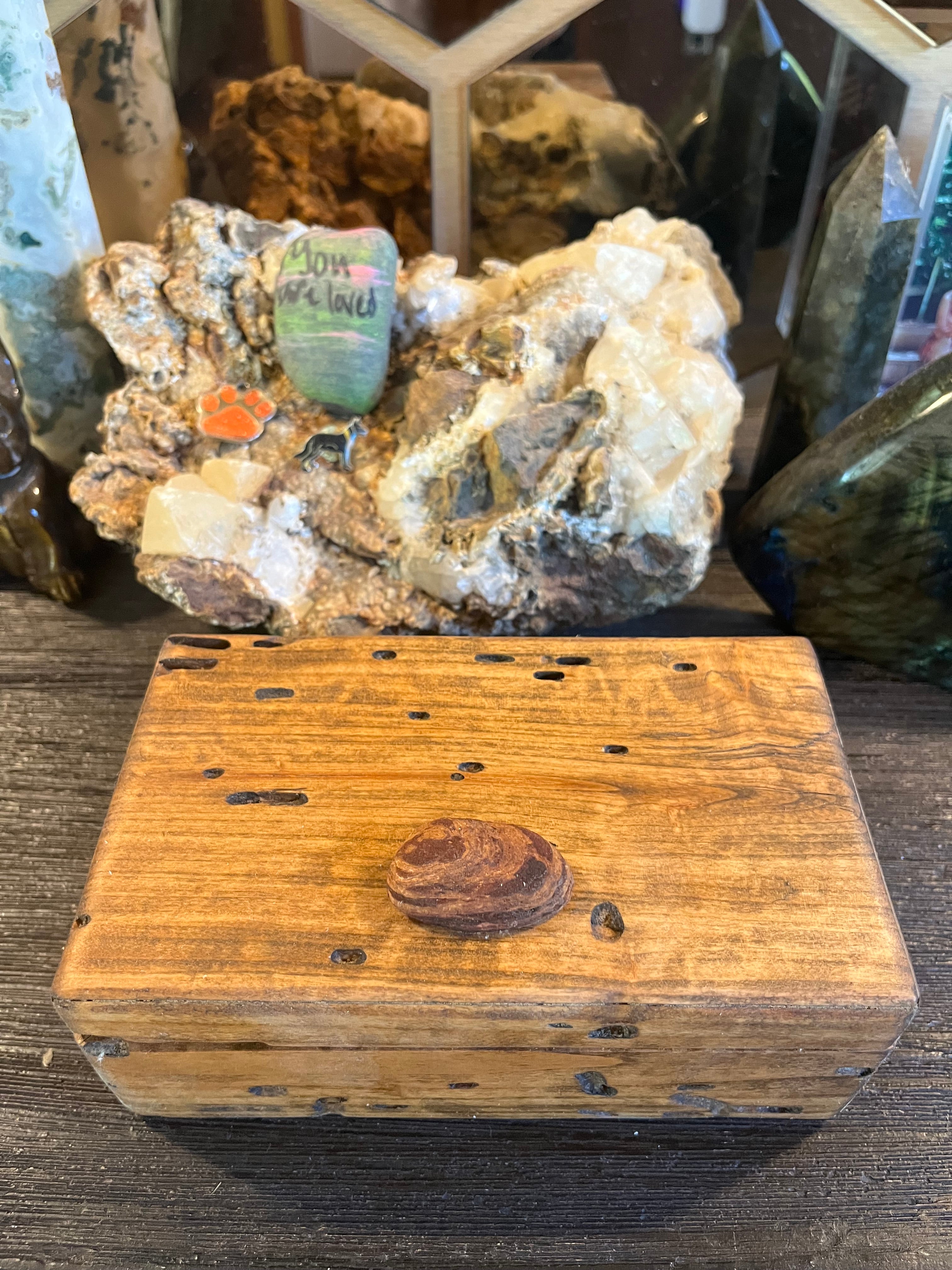 A rustic wooden keepsake box with a lid, featuring a small oval piece of Oregon driftwood on the center and several small cutouts on the lid, displayed on a dark wooden surface with crystals and minerals in the background.