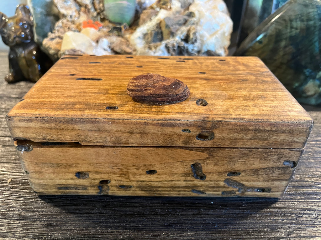 A rustic rectangular wooden box with a hinged lid, featuring natural wood grain, dark knots, and a small oval wooden knob on top made from Oregon cost driftwood, set on a dark wooden surface.