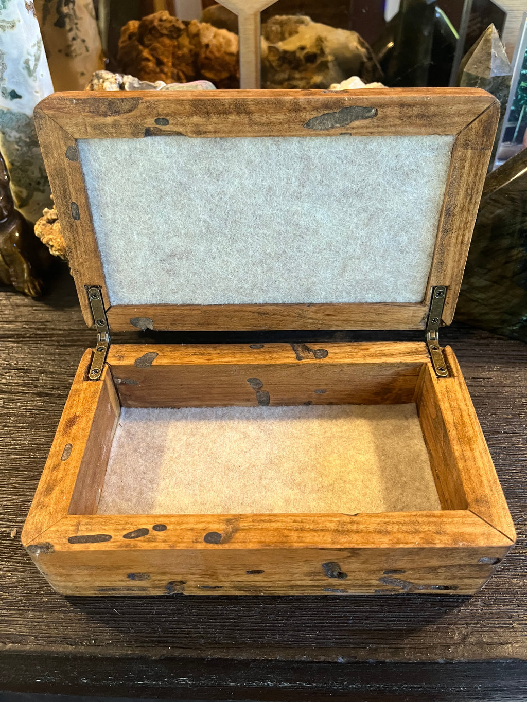 A rustic rectangular wooden box with an open hinged lid, lined with light beige felt on the inside and top panel, displayed on a dark wooden surface. The wood has very interesting patterns due to bug damage.
