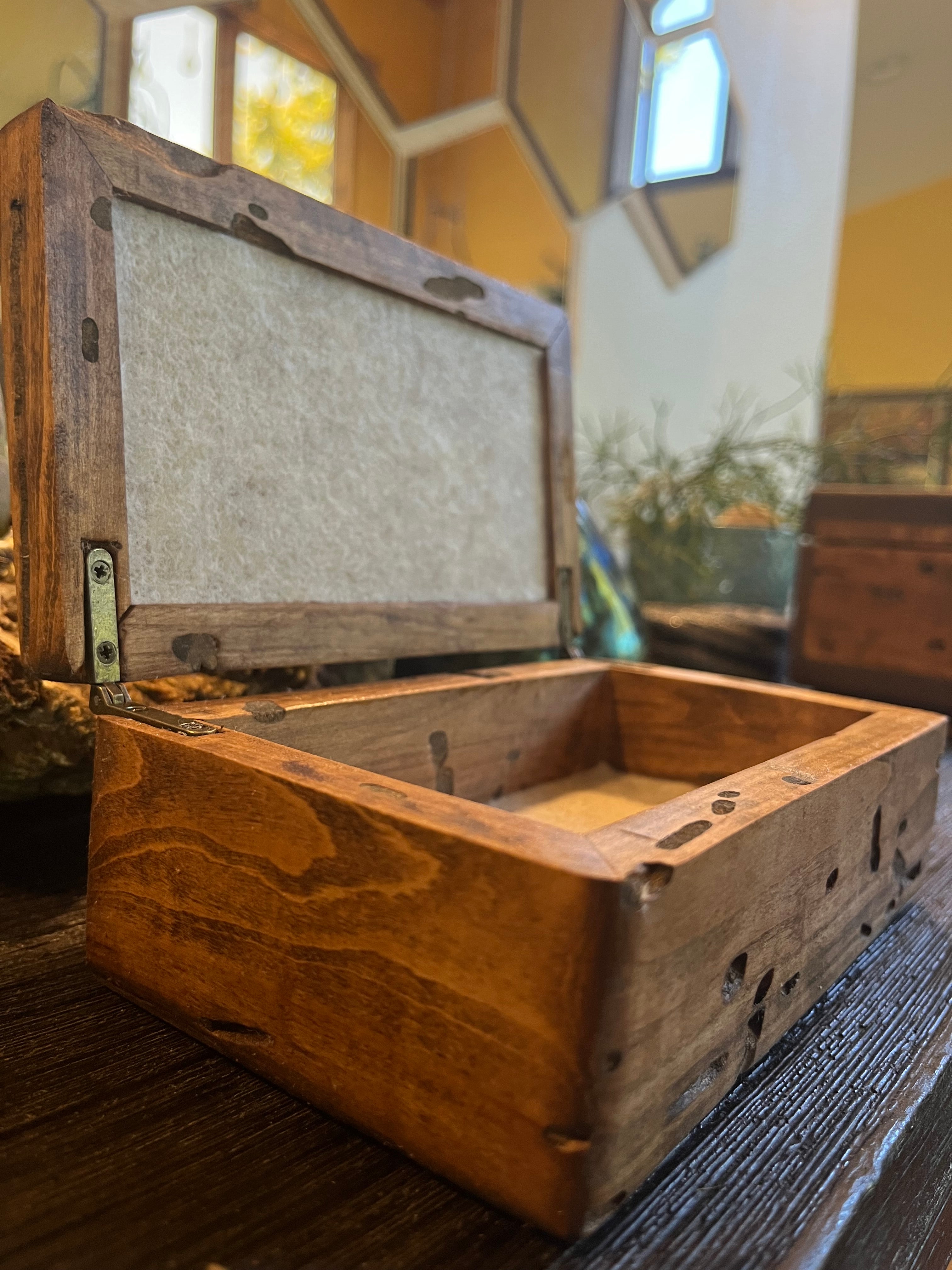 A rustic wooden keepsake box with an open hinged lid and a light beige felt lining, shown on a dark wooden table in a warm, softly blurred indoor setting. Made from locally grown Ash wood