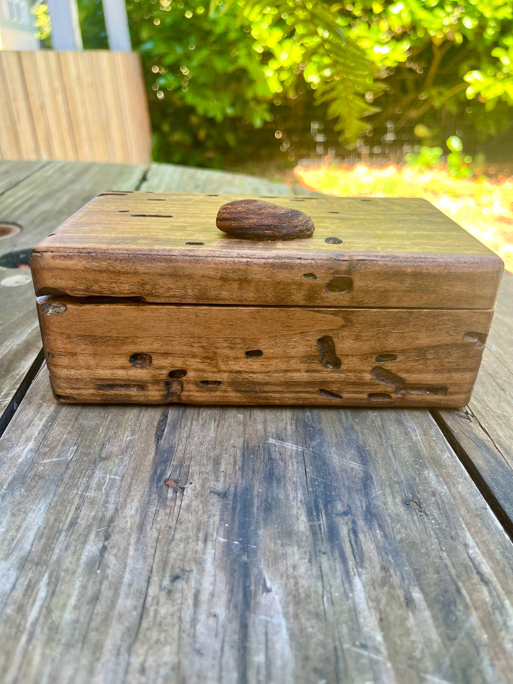 A rustic rectangular wooden box with a small oval wooden lid knob, resting on a weathered wooden table with a blurred garden background.