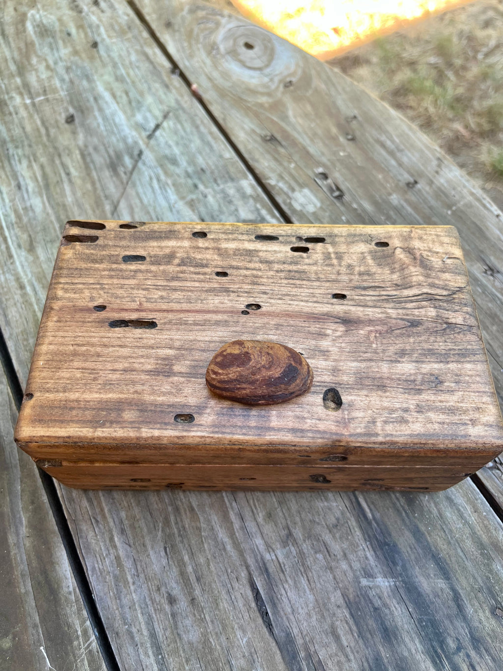 A rectangular wooden box with a hinged lid, featuring multiple small bug holes along the top surface and a small oval piece of Oegon beach wood on the top, resting on a weathered wooden deck.