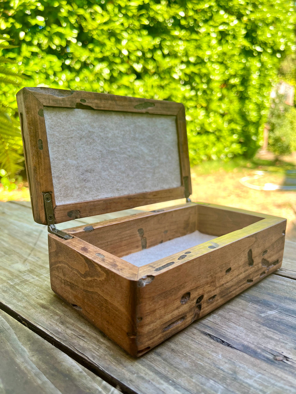 A rustic wooden keepsake box with an open hinged lid, lined with light beige felt, resting on a weathered wooden table with a blurred green garden background. very unique grain patterns and bug marks.
