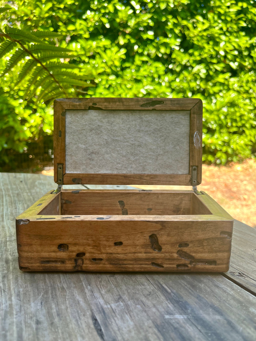 A rustic wooden keepsake box made from locally grown Ash wood with an open lid, resting on a weathered wooden table with a lush green garden background. lined top and bottom inside with soft gray felt. brass hinges hold the lid