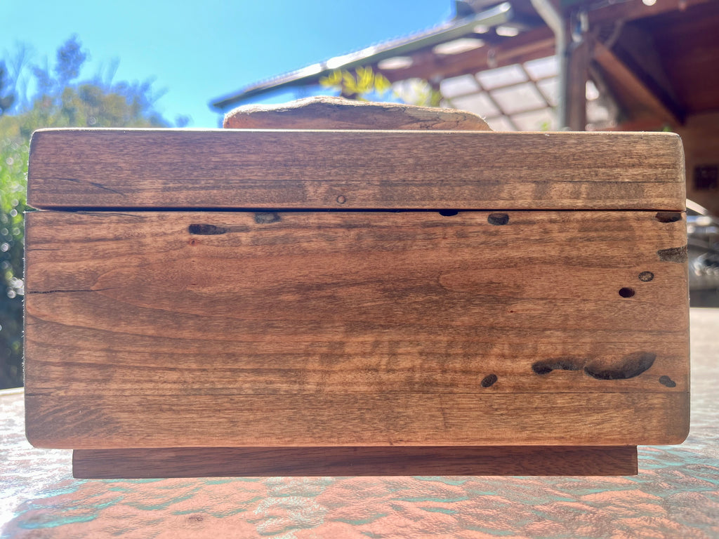 A rectangular wooden box with a hinged lid, made from weathered brown wood with visible grain, knots, and insect holes, sitting outdoors on a patterned surface.