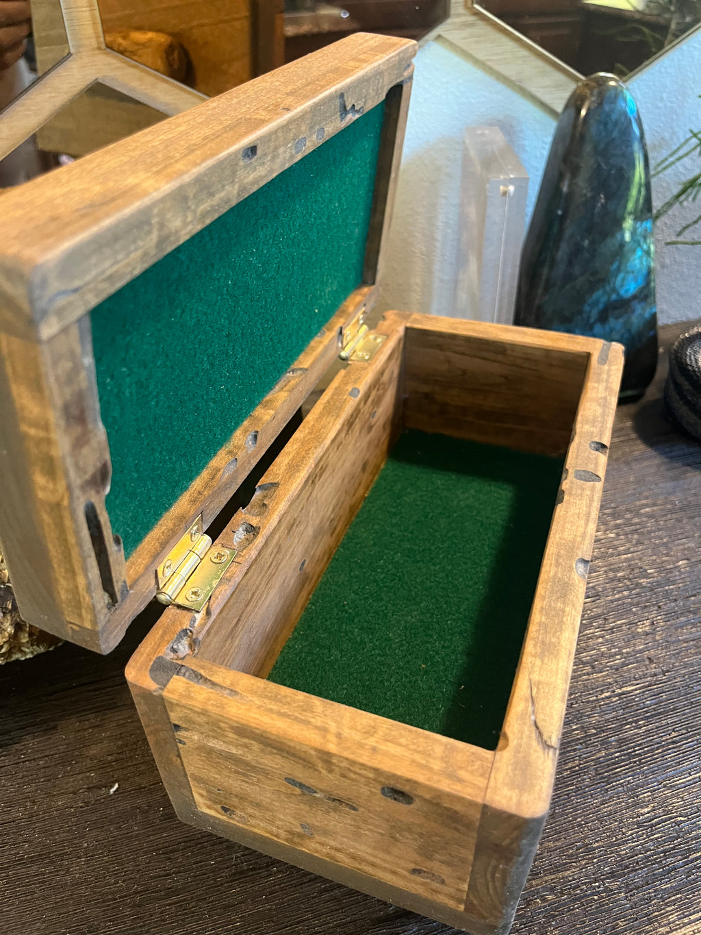 A rustic wooden jewelry box with an open hinged lid, lined with green velvet inside, resting on a dark wooden table. Locally grown wood with a lid pull from the Oregon coast