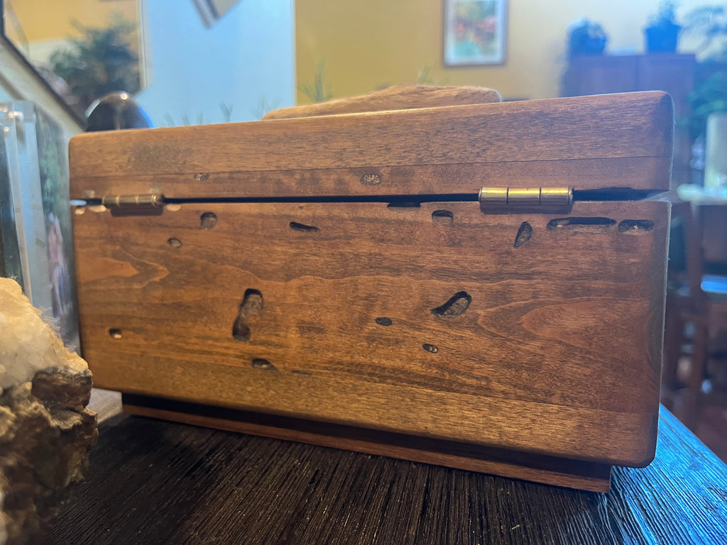 A rectangular wooden keepsake box with a hinged lid and brass-tone hinges, photographed on a dark tabletop in a real indoor setting with blurred furniture and decor in the background. Made from Locally grown Ash wood