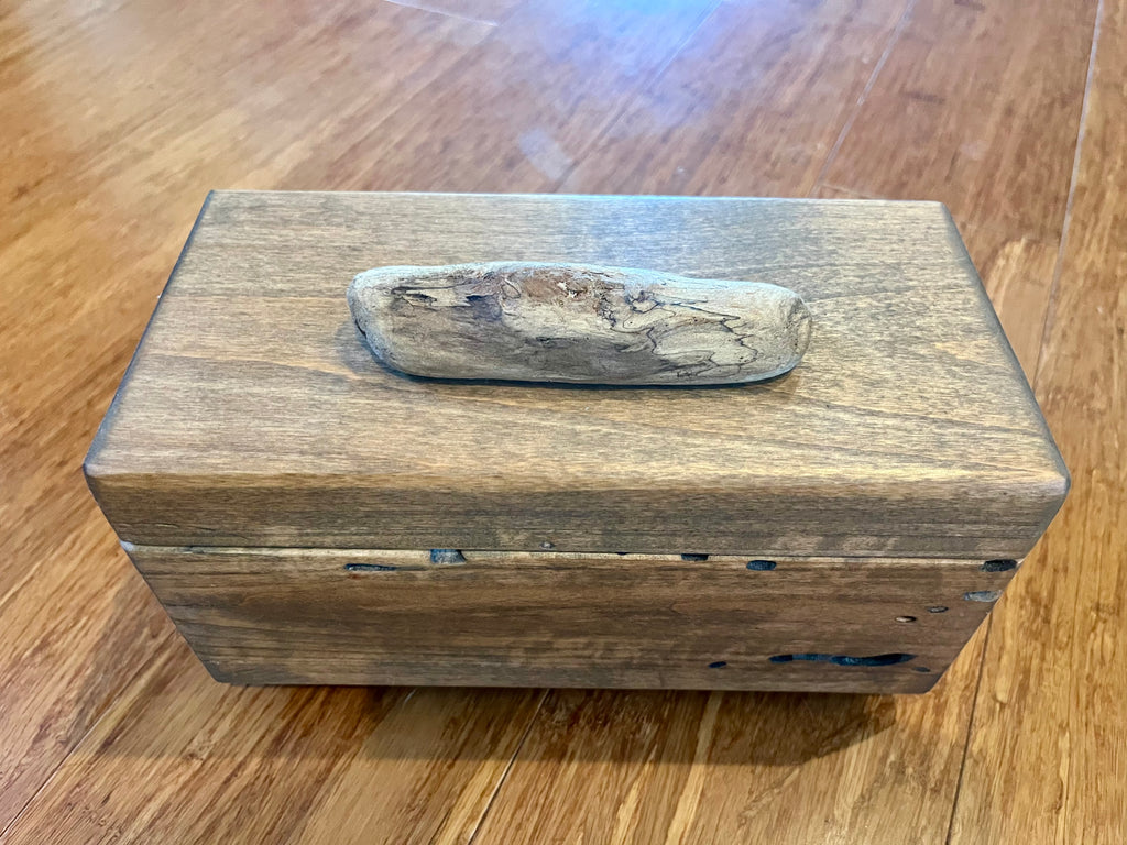 A rectangular wooden box made from locally grown Ash wood with a weathered driftwood handle on top, resting on a polished wooden floor. Finished with Tung Oil. 