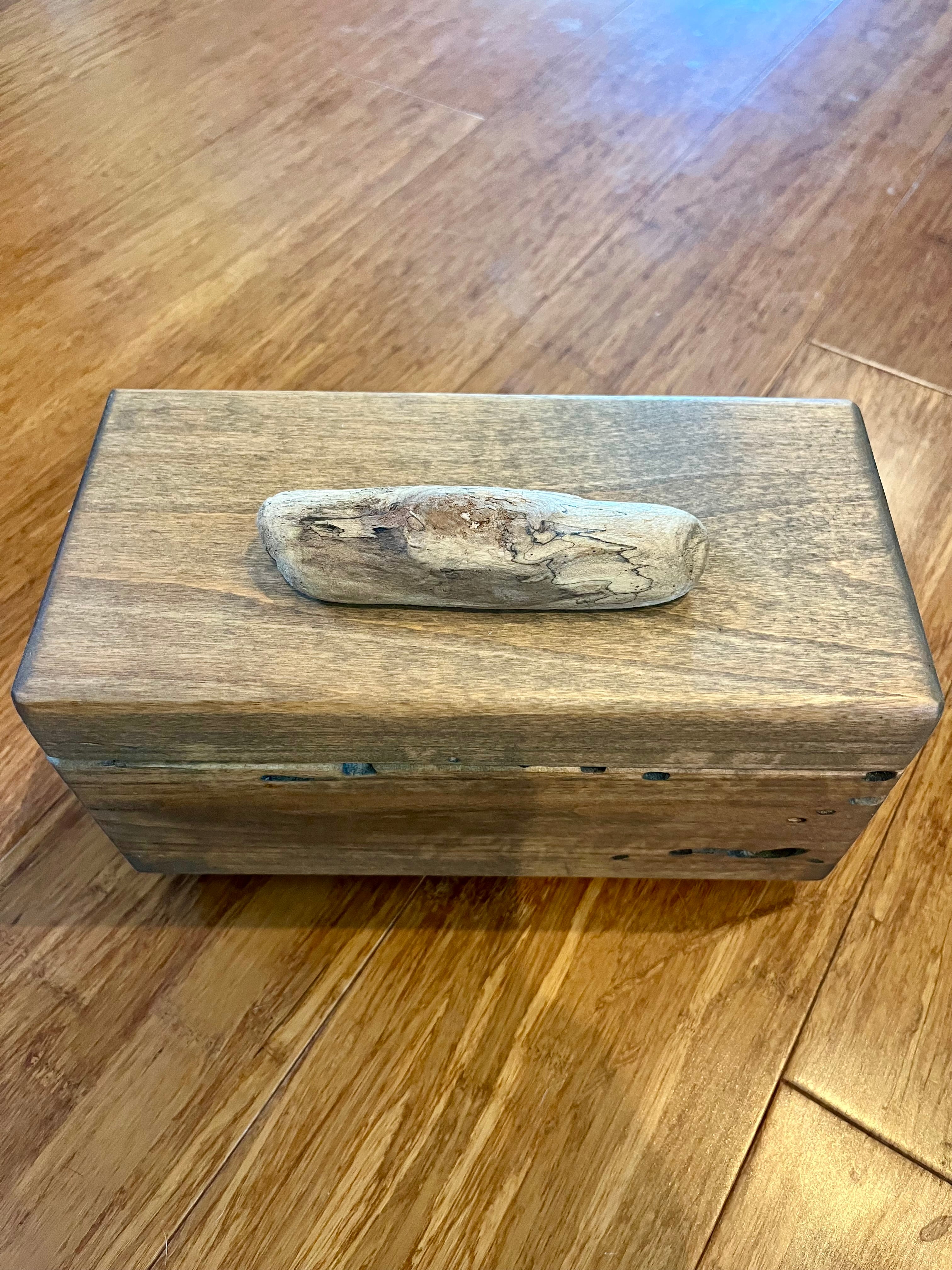 A rectangular Ash wood storage box with a rustic finish and a natural driftwood handle from the Oregon coast on top, resting on a polished wooden floor. The lid has brass hinges. 