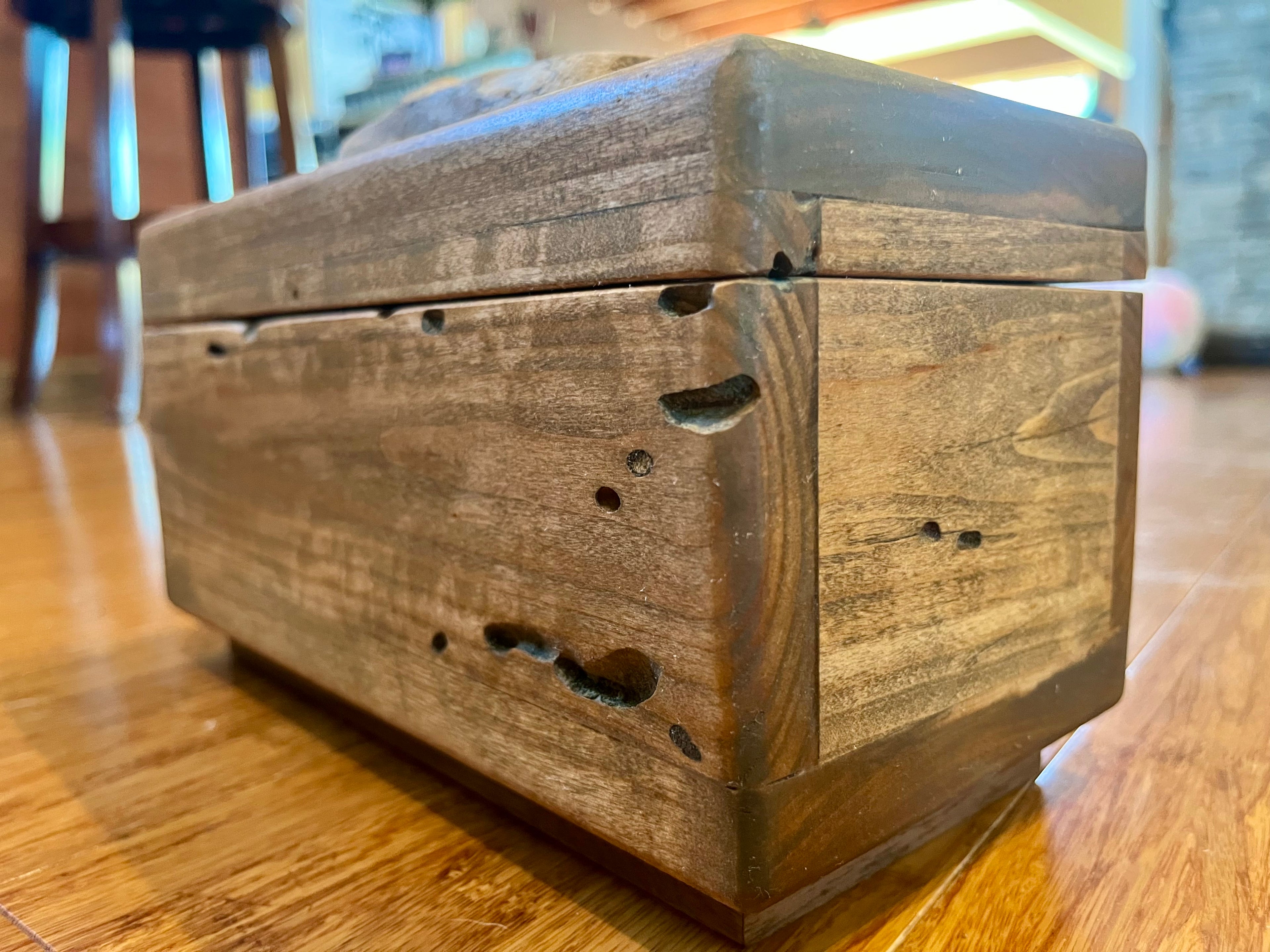 A rustic Ash wood storage box with a hinged lid, featuring prominent natural wood grain, knots, and rough edges, resting on a polished wooden floor.